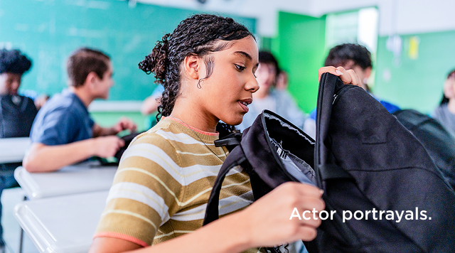 Teenage girl in a striped shirt putting a black jacket and bag into her backpack in a classroom.