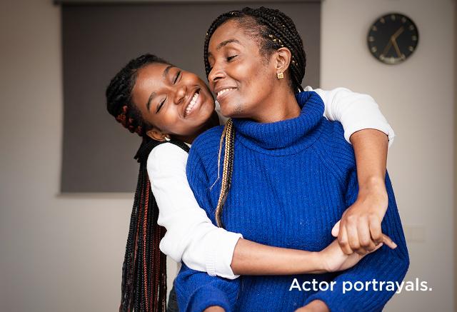 Smiling younger woman hugging a smiling older woman from behind in a kitchen.